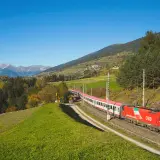 Roter ÖBB-Zug fährt durch eine grüne Alpenlandschaft. Die Strecke von Ljubljana nach Zagreb ist malerisch.