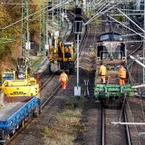 Arbeiter in orangefarbenen Warnwesten inspizieren Gleise neben Baumaschinen und Zügen. Die Szene zeigt die Infrastruktur der serbischen Eisenbahnen im Herbst.