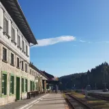 Der **Bayerisch Eisenstein Bahnhof** mit seinem historischen Gebäude und den grünen Fensterrahmen, umgeben von einer Waldkulisse unter blauem Himmel.