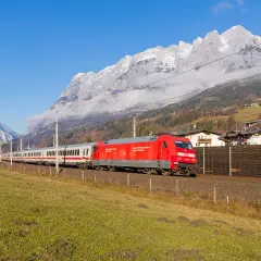 Ein roter Eurocity-Zug fährt durch eine grüne Wiese, im Hintergrund schneebedeckte Berge und ein Dorf.