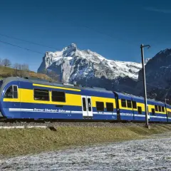 Blauer und gelber Zug der Berner Oberland-Bahn fährt durch eine verschneite Landschaft, mit schneebedeckten Bergen im Hintergrund. Die Fahrt von Interlaken nach Lauterbrunnen beginnt.