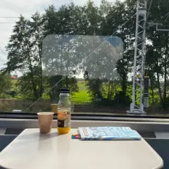 Blick aus dem Zugfenster auf die vorbeiziehende Landschaft. Auf dem Tisch stehen ein Becher, eine Saftflasche und ein Magazin. Die Reise von Warschau nach Berlin beginnt.