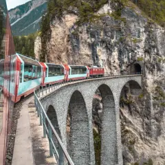Roter Zug fährt über eine Steinbogenbrücke in den Schweizer Alpen. Perfekt für eine Schweiz Rundreise Zug.