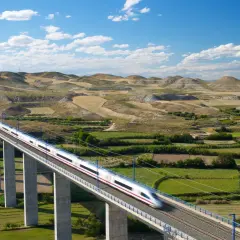 Schneller Zug auf einer Brücke in Spanien, der durch eine hügelige Landschaft mit Windrädern fährt.
