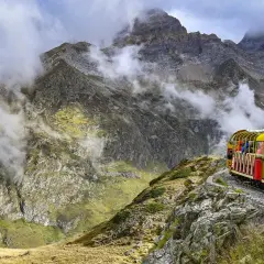 Gelber Zug fährt durch neblige Berglandschaft. Passagiere genießen die Aussicht auf steile Felsen und grüne Hänge.