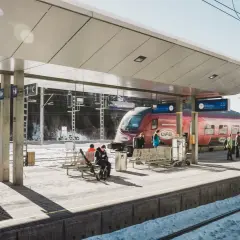 Winterliche Szene am **Lech Bahnhof**. Ein roter Zug steht bereit, während Reisende auf dem verschneiten Bahnsteig warten.