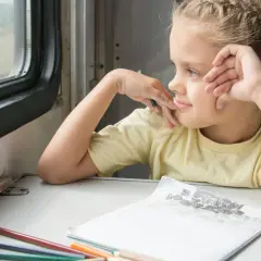 Ein Mädchen mit Zöpfen blickt verträumt aus dem Zugfenster. Buntstifte und eine Zeichnung liegen bereit für eine gute Zugfahrt.