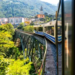 Mit dem Zug in die Toskana: Eine malerische Fahrt über eine Eisenbahnbrücke, vorbei an grünen Hügeln und einem charmanten Dorf mit Kirchturm.
