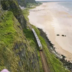 Ein Zug fährt entlang der Küste von **Railway Ireland**. Grüne Hänge stürzen zum Strand ab, das Meer glitzert in der Ferne.