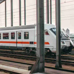 Moderner weißer IC-Zug mit orangefarbenen Streifen fährt auf Gleisen über eine Brücke. Im Hintergrund ist eine Stadt zu sehen.