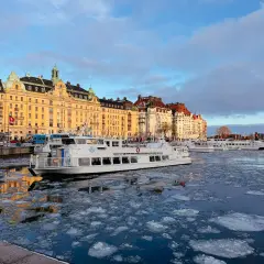 Eisbedeckter Kanal in Stockholm mit einem weißen Schiff, das an einer Promenade mit historischen Gebäuden vorbeifährt. Ein blauer Bus, der an einen schwedischen Zug erinnert, ist im Hintergrund zu sehen.