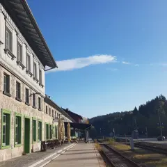 Der **Bayerisch Eisenstein Bahnhof** mit seinem historischen Gebäude und den grünen Fensterrahmen, umgeben von einer Waldkulisse unter blauem Himmel.
