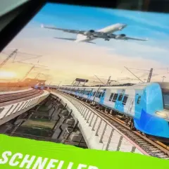 Blauer Hochgeschwindigkeitszug fährt auf erhöhter Strecke, während ein Flugzeug am Himmel schwebt. Eine Anzeige wirbt für "Zug statt Flug".
