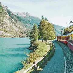Ein roter und gelber Fernzug fährt entlang eines türkisfarbenen Sees und majestätischer Berge.