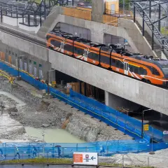 Moderner orange-schwarzer estonia train fährt auf einer erhöhten Strecke, während unten Bauarbeiten mit Baggern und blauen Absperrungen stattfinden.