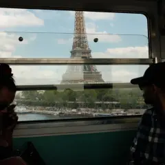 Blick aus dem Zugfenster auf den Eiffelturm. Zwei Reisende im Vordergrund, im Hintergrund die Seine und ein pariser Bahnhof.