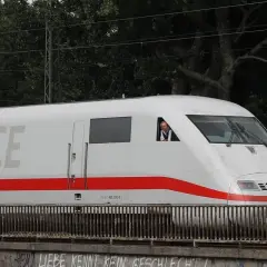 Ein Mann im Führerhaus eines weißen ICE-Zuges mit rotem Streifen. Der ICE-Service fährt auf einer Brücke.