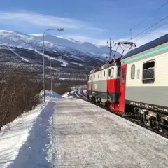 Der Zug von Stockholm nach Kiruna fährt durch eine verschneite Winterlandschaft mit schneebedeckten Bergen im Hintergrund.