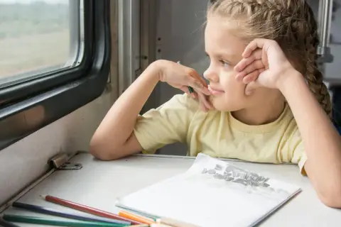 Ein Mädchen mit Zöpfen blickt verträumt aus dem Zugfenster. Buntstifte und eine Zeichnung liegen bereit für eine gute Zugfahrt.