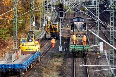 Arbeiter in orangefarbenen Warnwesten inspizieren Gleise neben Baumaschinen und Zügen. Die Szene zeigt die Infrastruktur der serbischen Eisenbahnen im Herbst.