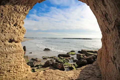Blick aus einer Höhle am Strand von Rom. Wellen brechen an Felsen.