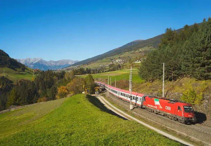 Roter ÖBB-Zug fährt durch eine grüne Alpenlandschaft. Die Strecke von Ljubljana nach Zagreb ist malerisch.
