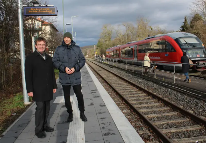 Zwei Männer stehen auf dem Bahnsteig des kleinsten Bahnhofs Deutschlands. Ein roter Zug der Westfrankenbahn wartet auf Fahrgäste.