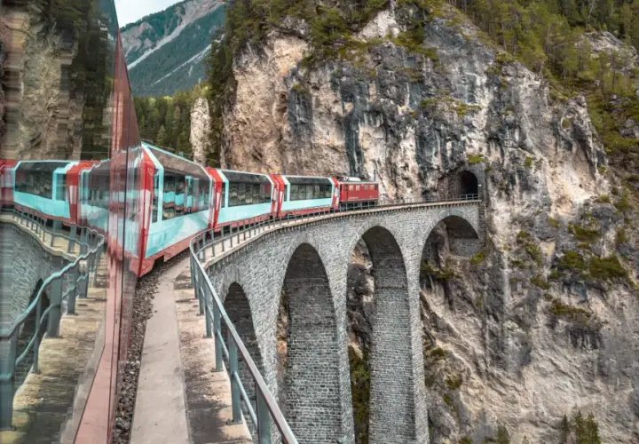 Roter Zug fährt über eine Steinbogenbrücke in den Schweizer Alpen. Perfekt für eine Schweiz Rundreise Zug.