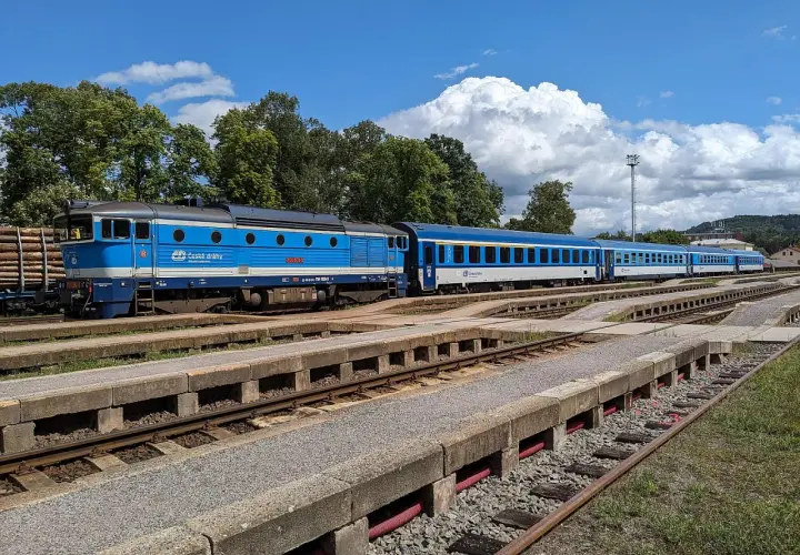 Blauer cz train mit mehreren Waggons steht auf Gleisen, umgeben von Bäumen und einem blauen Himmel mit Wolken.