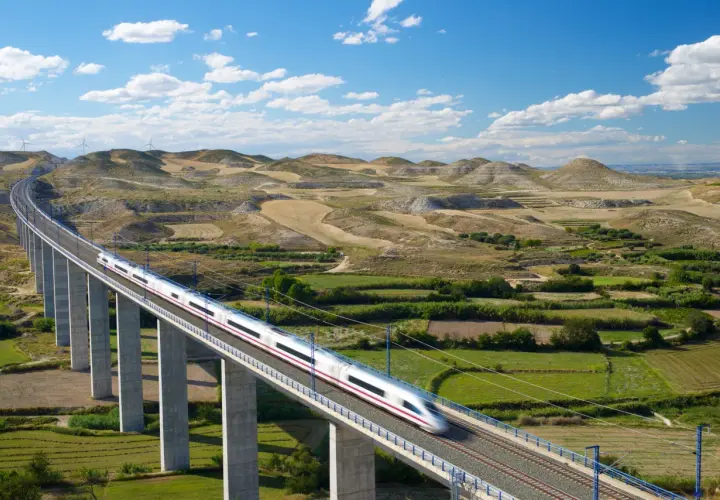 Schneller Zug auf einer Brücke in Spanien, der durch eine hügelige Landschaft mit Windrädern fährt.
