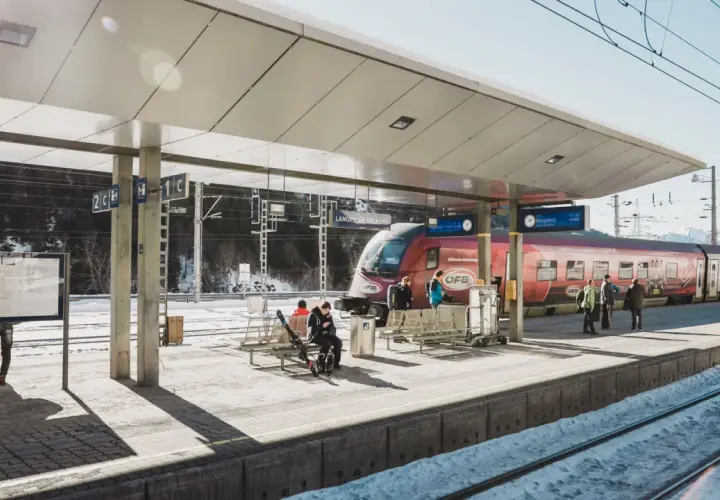 Winterliche Szene am **Lech Bahnhof**. Ein roter Zug steht bereit, während Reisende auf dem verschneiten Bahnsteig warten.