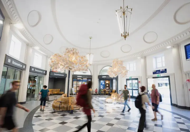 Menschen eilen durch den hellen, modernen **Coburger Bahnhof** mit seinen auffälligen Lampen und Sitzgelegenheiten.