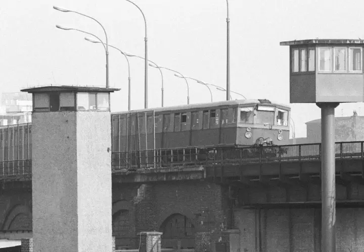 Eine Bahn fährt über eine Brücke, vorbei an Grenztürmen. Ein Symbol für die Zeit vor dem Mauerfall.