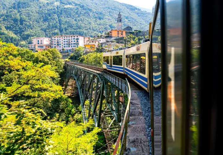 Mit dem Zug in die Toskana: Eine malerische Fahrt über eine Eisenbahnbrücke, vorbei an grünen Hügeln und einem charmanten Dorf mit Kirchturm.