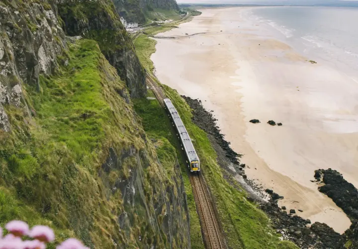 Ein Zug fährt entlang der Küste von **Railway Ireland**. Grüne Hänge stürzen zum Strand ab, das Meer glitzert in der Ferne.