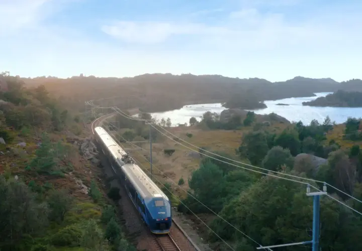 Ein blauer Zug fährt auf der Strecke Oslo-Stavanger durch eine hügelige, grüne Landschaft mit Seen und Felsen.