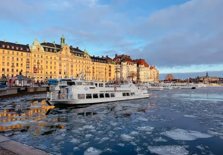 Eisbedeckter Kanal in Stockholm mit einem weißen Schiff, das an einer Promenade mit historischen Gebäuden vorbeifährt. Ein blauer Bus, der an einen schwedischen Zug erinnert, ist im Hintergrund zu sehen.