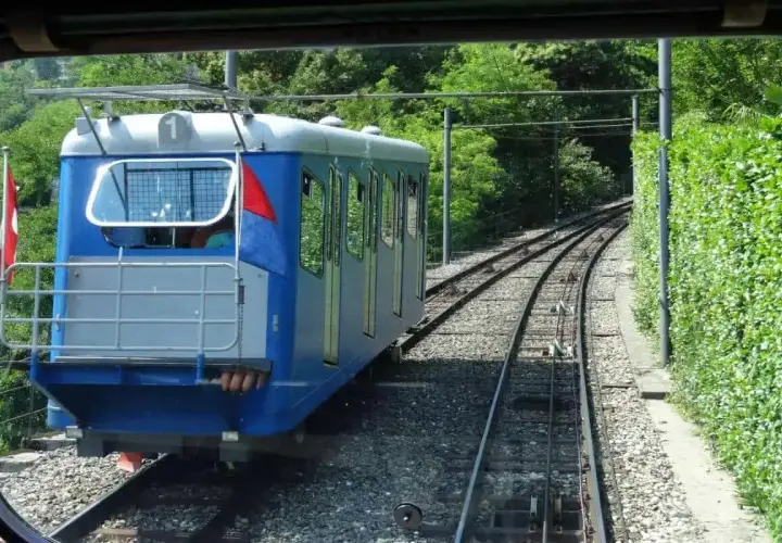Mit dem Zug zum Lago Maggiore: Eine blaue Standseilbahn fährt auf Schienen durch üppiges Grün.