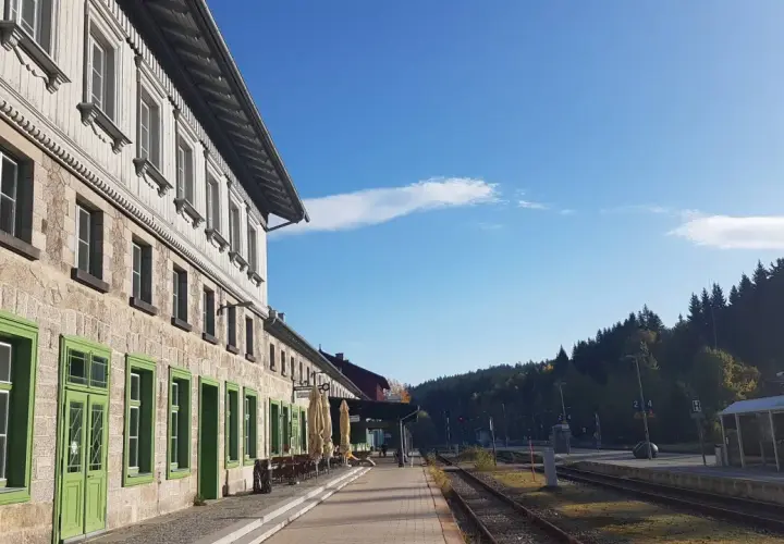 Der **Bayerisch Eisenstein Bahnhof** mit seinem historischen Gebäude und den grünen Fensterrahmen, umgeben von einer Waldkulisse unter blauem Himmel.