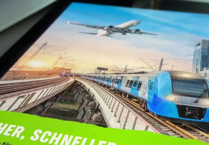 Blauer Hochgeschwindigkeitszug fährt auf erhöhter Strecke, während ein Flugzeug am Himmel schwebt. Eine Anzeige wirbt für "Zug statt Flug".