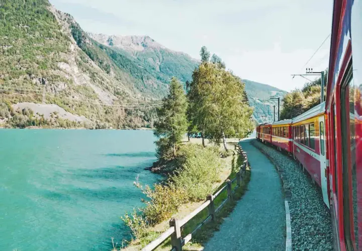 Ein roter und gelber Fernzug fährt entlang eines türkisfarbenen Sees und majestätischer Berge.