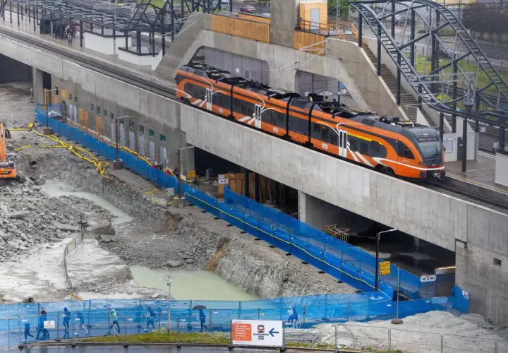 Moderner orange-schwarzer estonia train fährt auf einer erhöhten Strecke, während unten Bauarbeiten mit Baggern und blauen Absperrungen stattfinden.