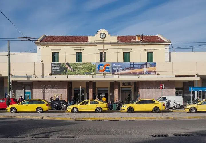 Gelbe Taxis warten vor dem Bahnhof Athen. Ein Schild verkündet "Wir transformieren unser Schienennetz, wir modernisieren Transporte".