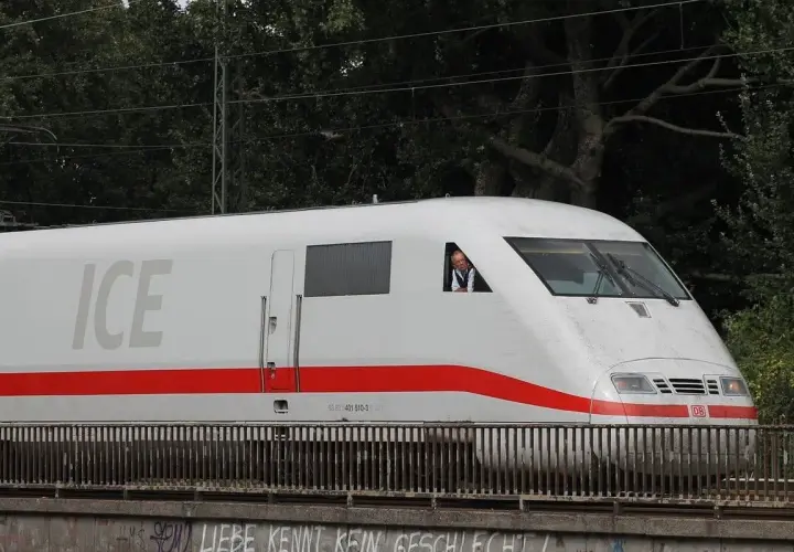 Ein Mann im Führerhaus eines weißen ICE-Zuges mit rotem Streifen. Der ICE-Service fährt auf einer Brücke.