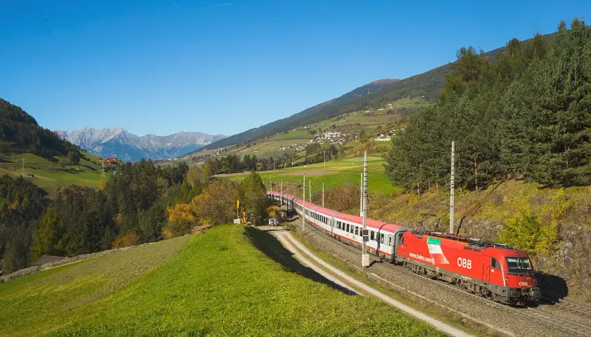 Roter ÖBB-Zug fährt durch eine grüne Alpenlandschaft. Die Strecke von Ljubljana nach Zagreb ist malerisch.