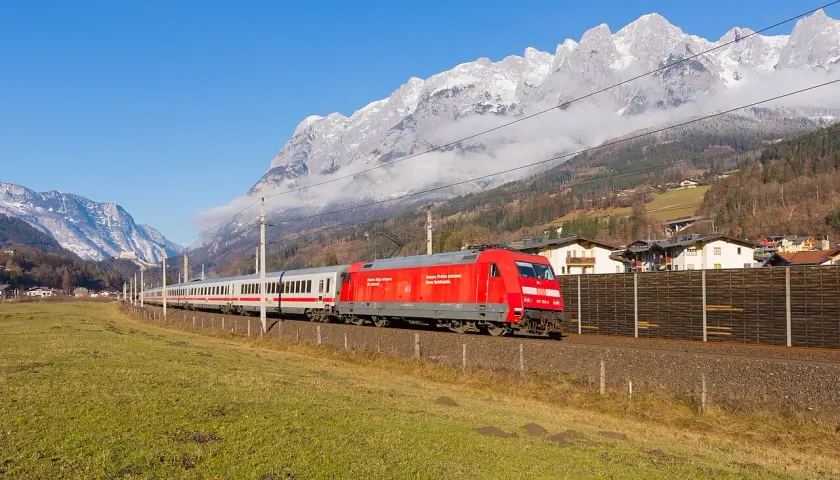 Ein roter Eurocity-Zug fährt durch eine grüne Wiese, im Hintergrund schneebedeckte Berge und ein Dorf.