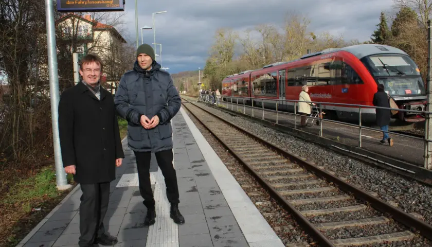 Zwei Männer stehen auf dem Bahnsteig des kleinsten Bahnhofs Deutschlands. Ein roter Zug der Westfrankenbahn wartet auf Fahrgäste.