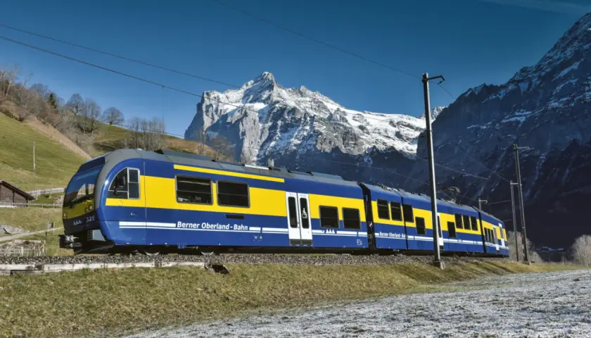 Blauer und gelber Zug der Berner Oberland-Bahn fährt durch eine verschneite Landschaft, mit schneebedeckten Bergen im Hintergrund. Die Fahrt von Interlaken nach Lauterbrunnen beginnt.