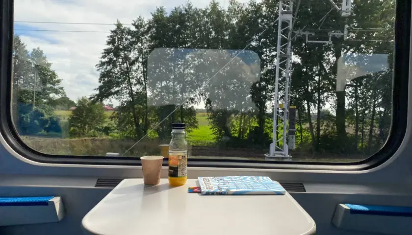 Blick aus dem Zugfenster auf die vorbeiziehende Landschaft. Auf dem Tisch stehen ein Becher, eine Saftflasche und ein Magazin. Die Reise von Warschau nach Berlin beginnt.