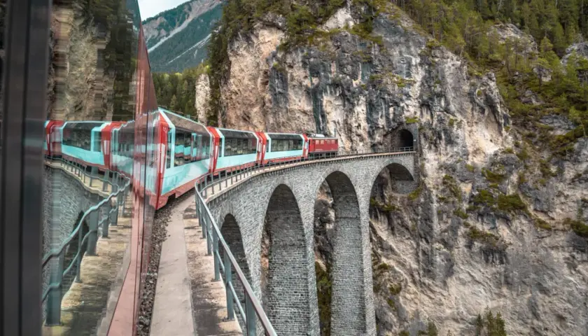 Roter Zug fährt über eine Steinbogenbrücke in den Schweizer Alpen. Perfekt für eine Schweiz Rundreise Zug.